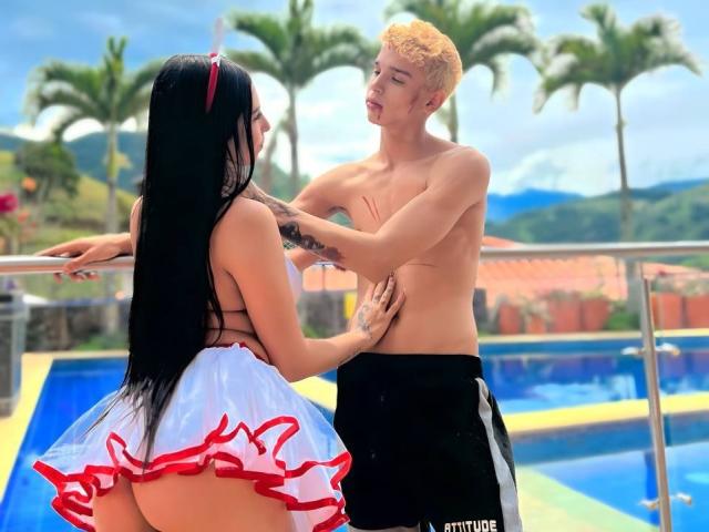 ArdienteCouple with long black hair and red headband, wearing a white and red frilled skirt, leans against a shirtless blond guy with black shorts by a pool.