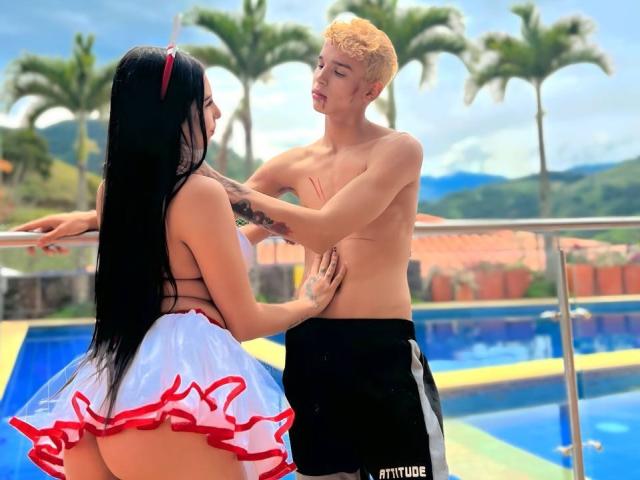 ArdienteCouple with black hair and red headband, wearing a white and red frilled skirt, facing a blonde, shirtless ArdienteCouple with black shorts, touching each other's torsos.