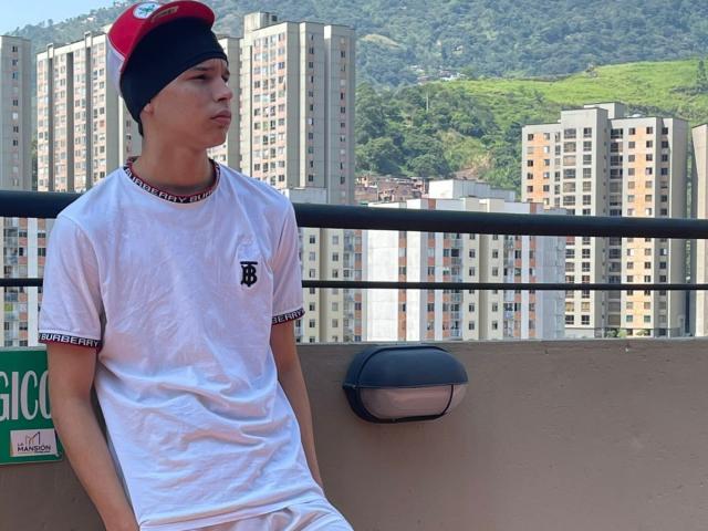 ArdienteCouple, wearing a red and black cap, white tee with black trim, sitting on a balcony, looking to the side, with tall buildings and green hills in the background.