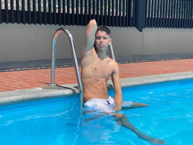 ArdienteCouple, shirtless with a slim, muscular build, sits in a blue pool, wearing white swim trunks, arm raised, hand on metal ladder, looking at the camera.