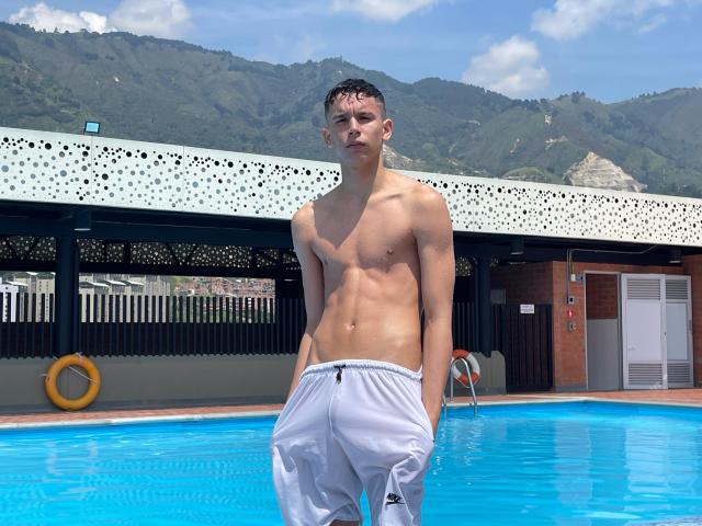 Shirtless ArdienteCouple with a slim, toned body, wet from the pool, wearing white pants, standing by the blue water, mountains in the background, and a perforated white roof above.