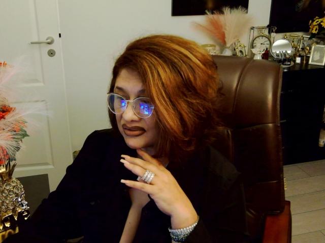 ClassybutNaughty has shoulder-length, wavy auburn hair. She's wearing clear, round glasses with blue light reflection and dark lipstick. Her right hand, with a silver ring, rests near her chin. She's in a brown leather chair, wearing a black jacket. The background has a white door, decorative silver and pink items, and a black shelf. The image has a casual, slightly messy vibe. The light is warm, highlighting her hair and glasses.