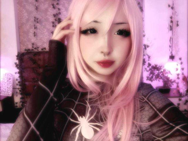 MillaHottLove69 con i capelli rosa, una maglia a rete nera con un disegno di ragno, un anello al naso e rossetto rosa, che si tiene i capelli. È carina, con grandi occhi e una luce rosa tenue..