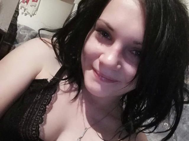 Elliefucku30-hot has pale skin, black wavy hair, and is smiling with her lips slightly parted. She's wearing a black lace top, showing her small tits and a silver necklace.