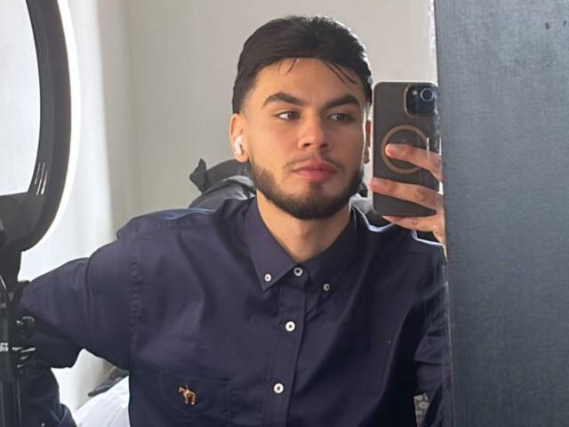 NastyX69 with dark hair, beard, and earbuds, wearing a navy shirt, taking a mirror selfie with a black phone, holding a ring light on the left.
