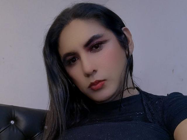 JaneStonesTs with dark hair, pink eyeshadow, red lipstick, and black sparkly top, looking down with a slightly sad expression, seated against a black leather tufted surface.