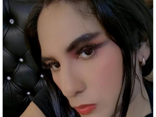 JaneStonesTs has dark, wavy hair, brown eyes, and red lipstick. Her face shows a slight blush and she's wearing subtle eyeliner. She's on a black tufted leather seat.