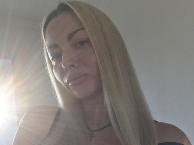 StefanieMargo with long blonde hair, light skin, and subtle makeup, sun flare in the left, slightly parted lips, wearing a black top, necklace, and confident expression.