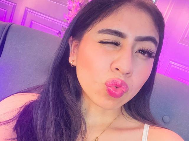 ZoeRedds with long black hair, winking, and puckering pink lipstick in a selfie, showing off her plump lips and gold necklace, sitting on a blue chair.