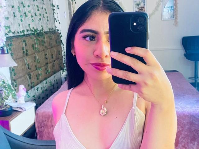ZoeRedds con il trucco rosa, che tiene in mano un telefono nero, indossa una canotta bianca, una collana con un ciondolo a forma di fiore, mostra il seno in un selfie, in una stanza illuminata di rosa..