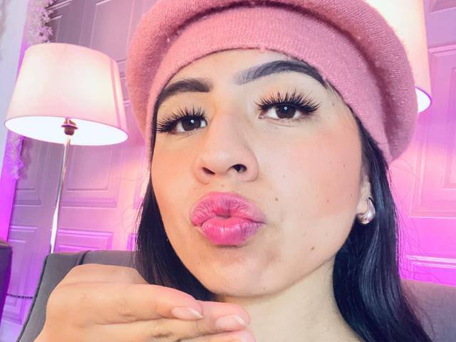 ZoeRedds with pink beanie, pouty pink lips, long eyelashes, brown eyes, and dark hair, making a kissy face, hand near mouth. Lamp in soft pink light.