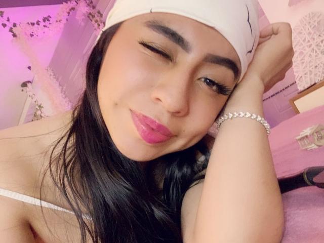 ZoeRedds, winking, with pink lipstick, black hair, white headband, and pink bracelet, resting her cheek on her hand, showing off her small tits and cute face.