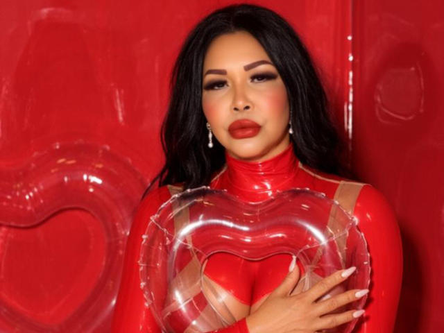 AngelinaDeleon with black hair, red lipstick, and red latex outfit holding a clear heart-shaped object over her tits. She has a confident, sexy look with her nails painted.