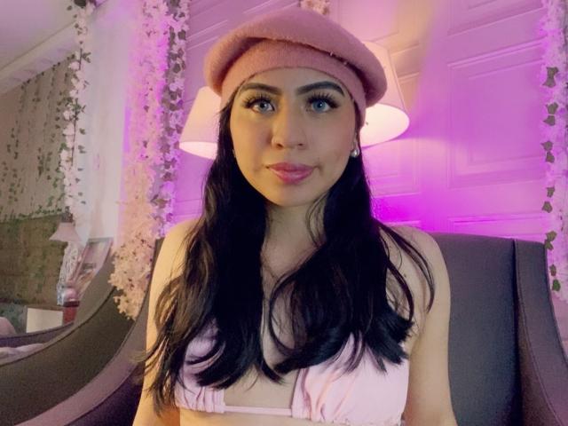 ZoeRedds with long black hair, pink beret, pink lipstick, and pink top, showing her small tits, sitting on a black chair, with purple and white lights in the background.