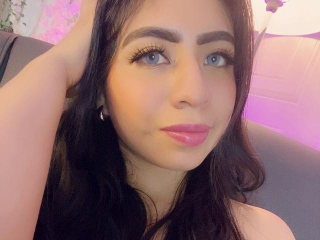 ZoeRedds has long black hair, blue eyes, and pink lipstick. She's touching her head with one hand, wearing pink earrings, and has thick, dark eyebrows. Her tits are not visible.