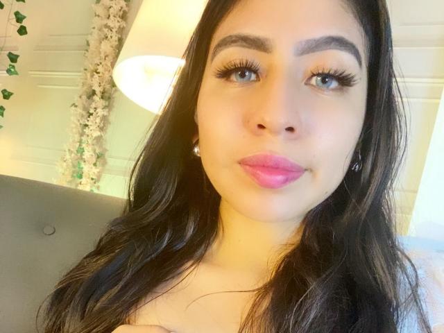 ZoeRedds tem cabelos longos e escuros, olhos azuis, sobrancelhas grossas, cílios longos, batom rosa e pequenos brincos. Ela está sorrindo e tem uma pele clara. Seus seios não estão visíveis..