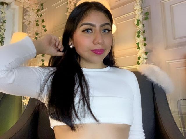 ZoeRedds with long black hair, wearing a white crop top, pink lipstick, and earrings, has her hand in her hair, showing her tits slightly. She's sitting in a chair.
