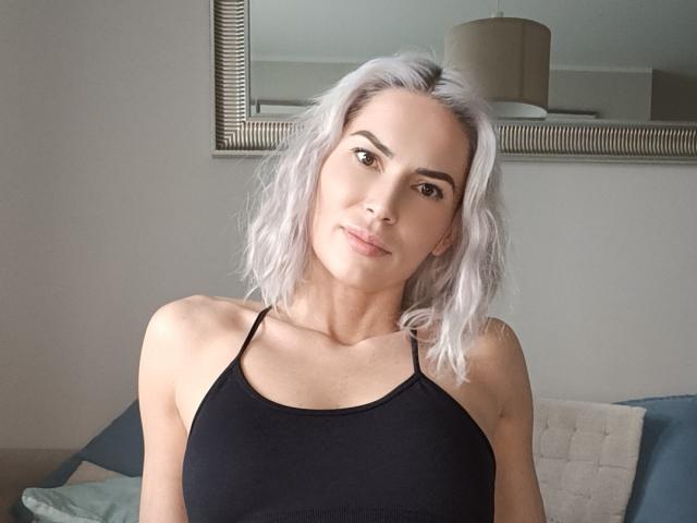 RowenaMinna with light purple hair, wearing a black spaghetti-strap top, showing small tits, smiling softly, brown eyes, light skin, in a sunlit room.