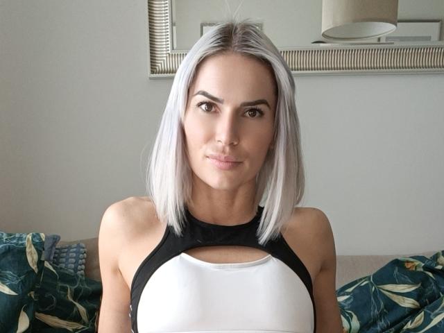 RowenaMinna with silver hair, black and white halter top, showing her small tits, sitting on a couch with green floral pillows, looking at the camera with a slight smile.