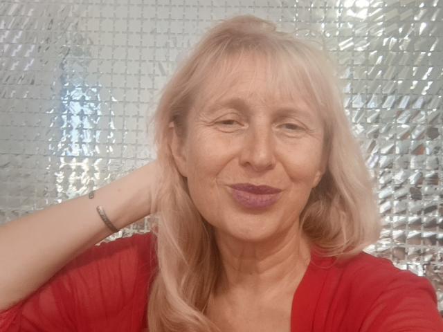 TrinaAdair has pale skin, blonde hair, and is smiling with purple lipstick. She's wearing a red shirt, has a bracelet on her right wrist, and is looking at the camera.