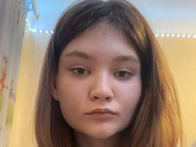 MagdaleneFein has brown hair, brown eyes, and a slightly pouty face with a small mole on her left cheek. Her eyebrows are thick, and she's looking straight at the camera.