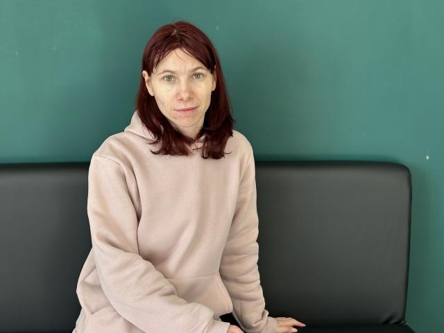 KateFire has pale skin, red hair, and wears a pink hoodie. She's sitting on a black bench against a green wall, with a slight smile on her face.