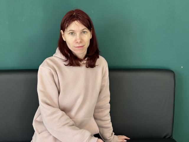 KateFire has pale skin, red hair, and green eyes, wearing a light pink hoodie, sitting on a black bench against a teal wall, with a subtle smile.