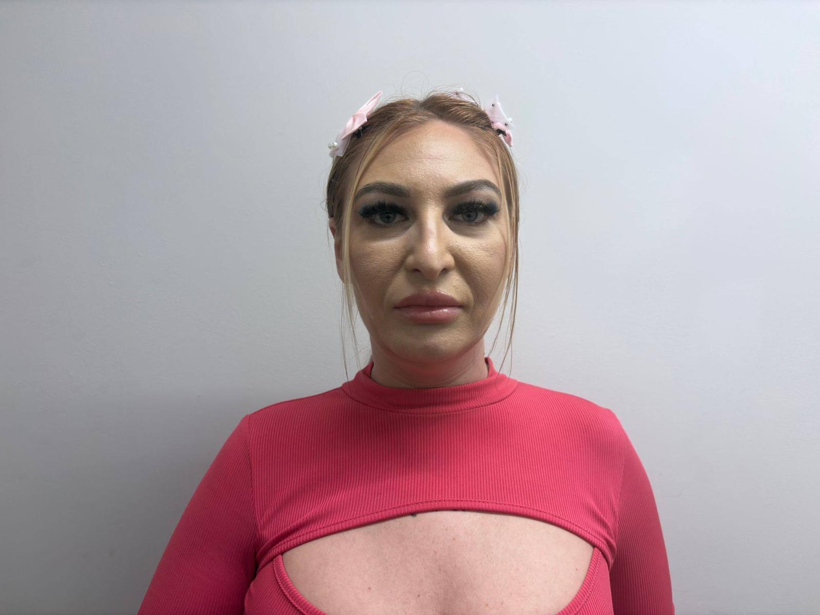 GraceQuenny with light brown hair, pink flowers in hair, heavy makeup, pink long-sleeve top with cutout showing her tits, neutral expression, standing against plain background.
