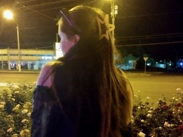 Drayana with long brown hair, a star hair clip, and a black off-shoulder top, standing by a flower bed at night, her back to the camera.