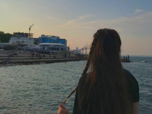 Drayana with long brown braids, facing waterfront, holding braid, back to camera, wearing black shirt, water in foreground, buildings and crane in background, sunset sky.