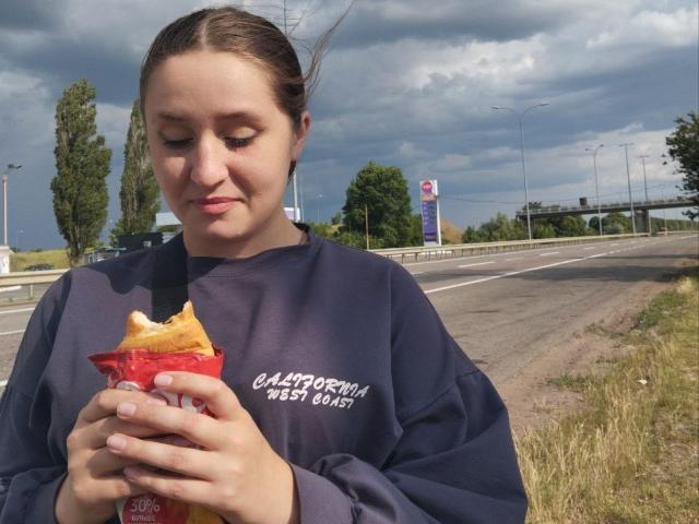 Drayana with brown hair in a dark sweatshirt eating a bag of chips on a road, holding it with both hands, under a cloudy sky, looking down.