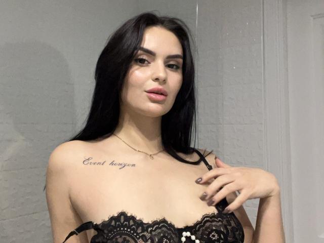 MilashaLovely with long black hair, wearing black lace lingerie, showing small tits, has tattoo on right chest, pink lipstick, and a gold necklace, with hand on chest.
