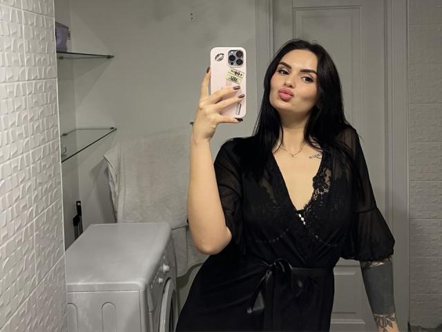 MilashaLovely with black hair, pouty lips, and tattoos, wearing a black sheer robe, taking a selfie in a mirror with a pink phone, showing her tits.