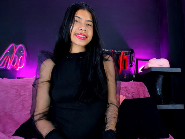 ChloeStone with long black hair, wearing a sheer black dress, black gloves, and a jeweled headband, smiling with red lipstick, sitting on a pink, furry couch.