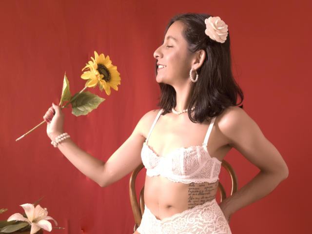 MiaAmoree, a woman with medium-length dark brown hair, wears a white lace bra and matching lace panties. She holds a sunflower in her right hand, eyes closed in a joyful expression. She has a tattoo on her right side, a pink flower in her hair, and wears a pearl necklace and bracelet. The image has a red background with a wooden chair and a lily in the bottom left. Her fair skin contrasts with the lace and flower. She has small to medium-sized breasts.