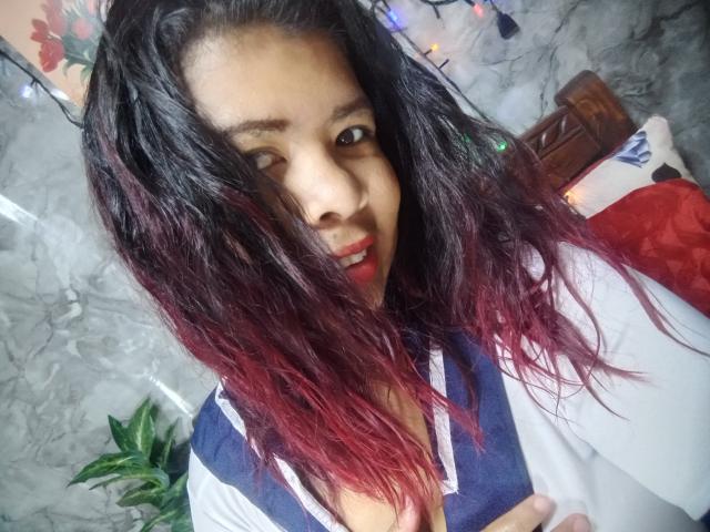 MariahSquirting69 with wet, dark red to black ombre hair, winking, and showing slight cleavage in a blue and white top, with a small green plant on the left.