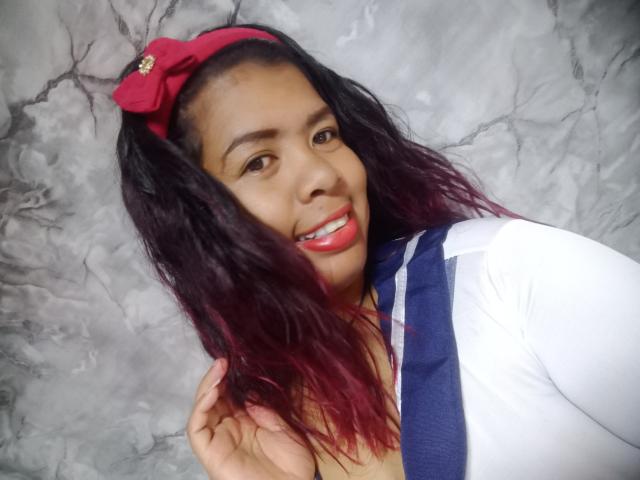 MariahSquirting69 tiene la piel de color marrón oscuro, lápiz labial rojo y cabello negro ondulado con reflejos rojos, lleva un lazo rojo, una blusa azul y blanca, está sonriendo y tocándose el cabello..