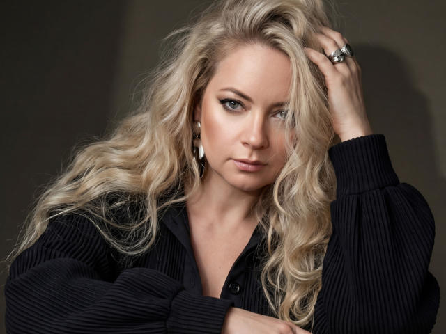 CharlotteKerti with long, wavy blonde hair, wearing a black ribbed shirt, hand in hair, blue eyes, serious expression, silver earrings, and a ring, looking directly at camera.