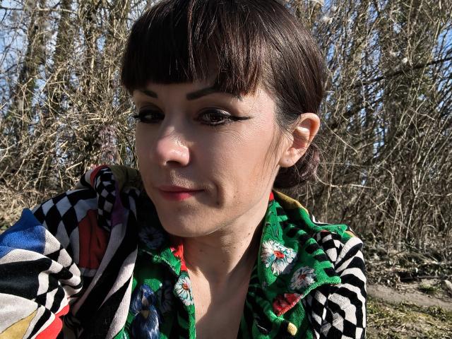 OrianaLaFrancaise has a light skin tone, dark brown, straight hair with blunt bangs, and wears bold, black, winged eyeliner. She is dressed in a colorful floral and geometric patterned shirt. Her makeup is subtle with a hint of pink lipstick. She is looking slightly to the left with a slight, closed-mouth smile. The background is a natural, leafless, outdoor setting with branches and a clear, blue sky. The lighting is bright and natural, highlighting her facial features and the intricate patterns of her shirt. The image is sharply focused.