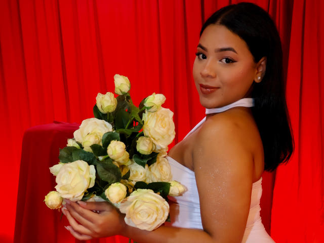 AuroraaRose with dark hair, wearing a white halter dress, holding a bouquet of white roses, smiling at the camera, red background, soft light, slightly glossy skin.