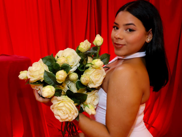 AuroraaRose with dark hair, holding a bouquet of white roses, wearing a white halter dress, smiling at the camera, red curtain in the background, looking sexy.