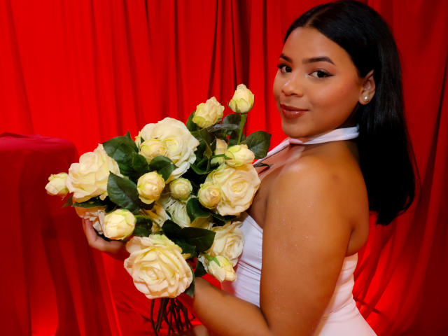 AuroraaRose with tan skin, black hair, white halter dress, holding a bouquet of yellow roses, smiling at the camera, red background, her tits slightly visible, looking sexy.