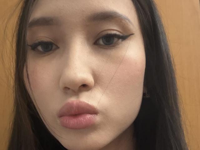 Close-up of AndrianaFive's face, center frame. She has light skin, full pink lips, and dark, slightly wavy hair. Her eyes are medium-sized with black, winged eyeliner that stands out. A thin, almost invisible hair is visible on her right cheek. The background is a simple, warm brown, slightly out of focus. The texture of her skin is smooth with a subtle blush on her cheeks. AndrianaFive's expression is neutral, with a slight pout. Her eyebrows are thin and dark.