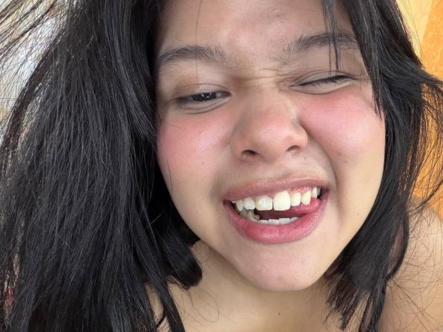 Mei69 tiene el cabello oscuro y despeinado, y un rostro ligeramente rojizo e hinchado, con la boca entreabierta mostrando los dientes. Está guiñando un ojo y parece molesta. Sus labios rosados están entreabiertos..