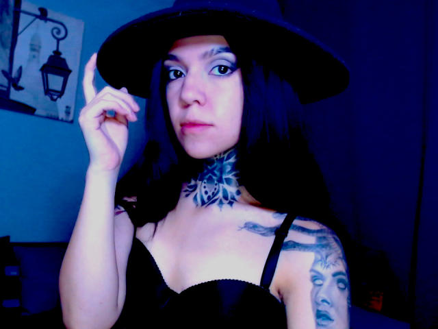 LannieGonzales with black hair, wearing a black hat, black bra, and tattoos, holding her hat, blue-lit, showing tattoos on neck and arm, with visible tits and a pretty face.