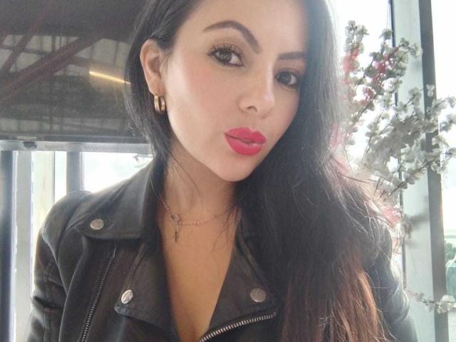 JakyKidman has long black hair, red lipstick, and is pouting with a black leather jacket. She's wearing gold earrings and a delicate necklace. Her tits are partially visible.