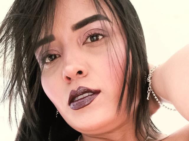 MelizaQueen has dark, glossy lipstick, thick eyebrows, and black hair with bangs. She's wearing a silver chain necklace and has a slightly open mouth. Her eyes are half-closed.