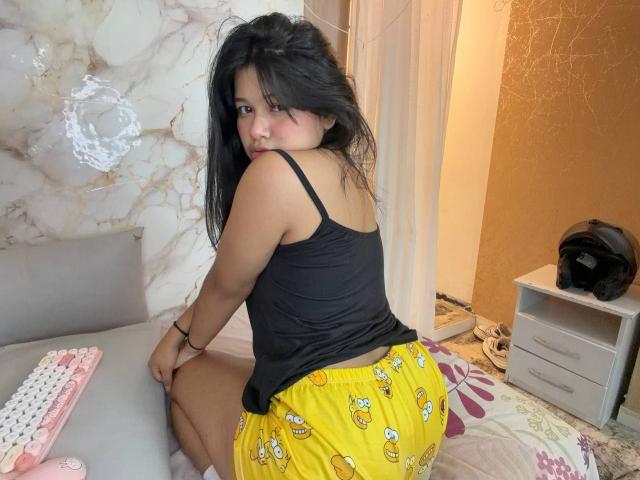 Mei69 vestindo uma camiseta preta e shorts de pijama amarelos com estampas de banana, sentada na cama, olhando para trás, mostrando o bumbum, cabelos pretos longos, pele morena, em um quarto..
