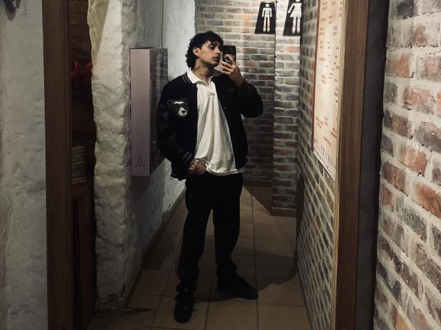 JayTaylor è in piedi davanti a uno specchio, in un autoritratto, indossando una giacca nera, una camicia bianca e pantaloni neri, con capelli ricci e scuri, tiene in mano un telefono, in un corridoio con pareti in mattoni..