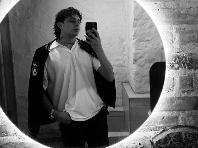 Autoportrait en noir et blanc de JayTaylor, avec des cheveux bouclés, portant une chemise polo blanche, une veste sombre et un bracelet, tenant un téléphone, debout devant un mur texturé..